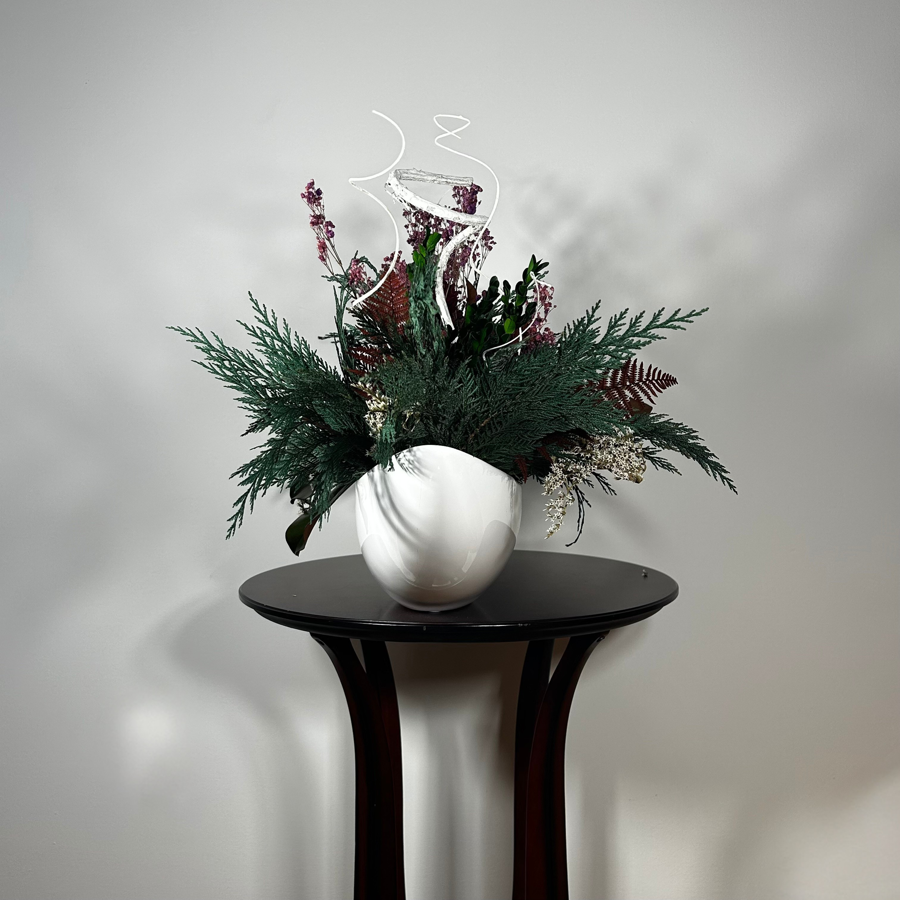 This dried flower centrepiece uses a low white lacquered vessel in a crescent shape. The foundation is a textural marvel, featuring the rich greens of preserved Cedar and Boxwood, contrasted by the striking texture of preserved Magnolia leaves and preserved burgundy Bracken. The dramatic florals include magnificent dried Dahlias in a deep crimson and elegant preserved Gypsophila dyed deep rose. Dried German Statice adds airy texture, and white curly ting ting and white wash curly vine complete the look.