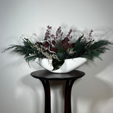 This dried flower centrepiece uses a low white lacquered vessel in a crescent shape. The foundation is a textural marvel, featuring the rich greens of preserved Cedar and Boxwood, contrasted by the striking texture of preserved Magnolia leaves and preserved burgundy Bracken. The dramatic florals include magnificent dried Dahlias in a deep crimson and elegant preserved Gypsophila dyed deep rose. Dried German Statice adds airy texture, and white curly ting ting and white wash curly vine complete the look.