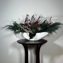 This dried flower centrepiece uses a low white lacquered vessel in a crescent shape. The foundation is a textural marvel, featuring the rich greens of preserved Cedar and Boxwood, contrasted by the striking texture of preserved Magnolia leaves and preserved burgundy Bracken. The dramatic florals include magnificent dried Dahlias in a deep crimson and elegant preserved Gypsophila dyed deep rose. Dried German Statice adds airy texture, and white curly ting ting and white wash curly vine complete the look.