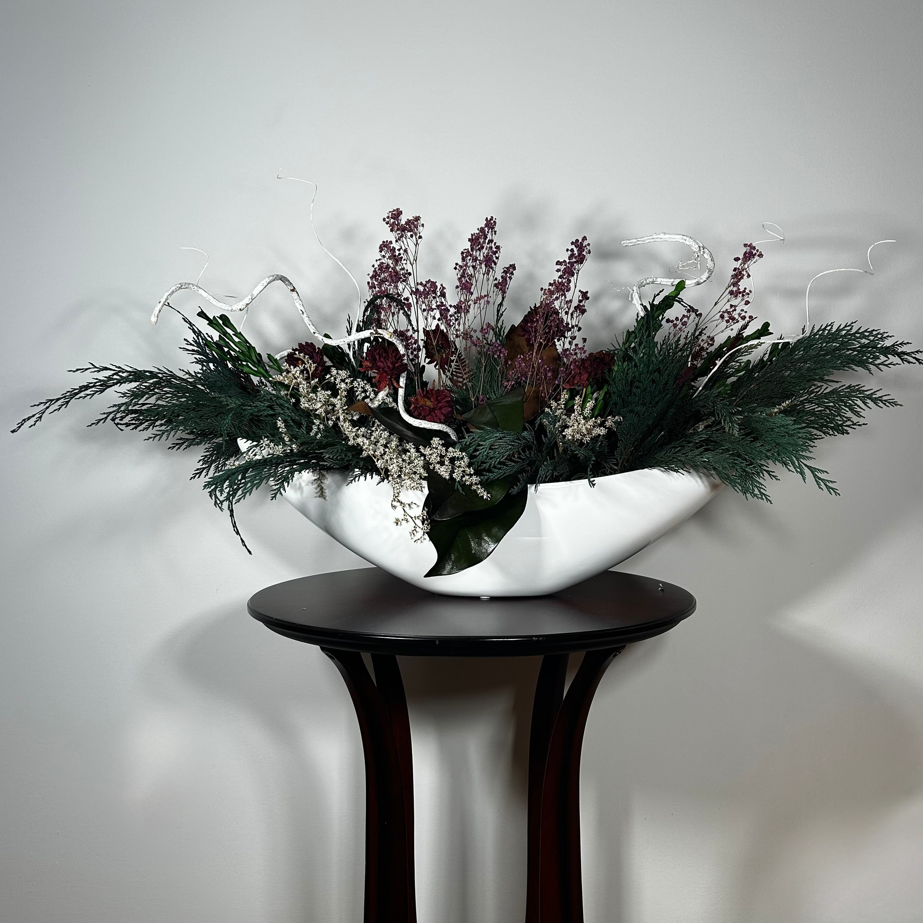 This dried flower centrepiece uses a low white lacquered vessel in a crescent shape. The foundation is a textural marvel, featuring the rich greens of preserved Cedar and Boxwood, contrasted by the striking texture of preserved Magnolia leaves and preserved burgundy Bracken. The dramatic florals include magnificent dried Dahlias in a deep crimson and elegant preserved Gypsophila dyed deep rose. Dried German Statice adds airy texture, and white curly ting ting and white wash curly vine complete the look.