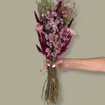 This striking, hand-tied bouquet is a harmonious blend of dried botanicals in deep red, soft pink, and natural oats. In this image a hand is holding the bouquet against a plain green background.
