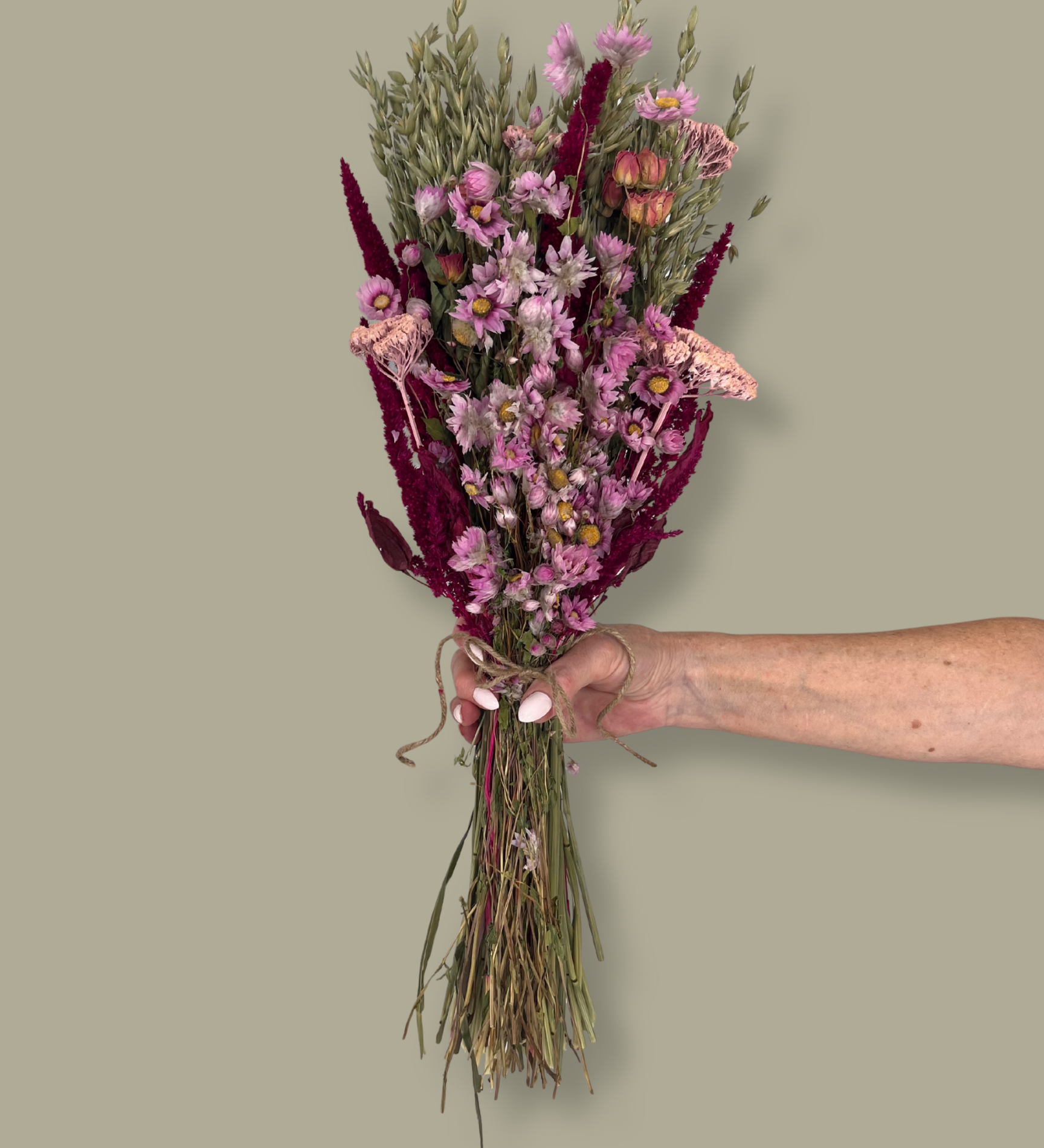 This striking, hand-tied bouquet is a harmonious blend of dried botanicals in deep red, soft pink, and natural oats. In this image a hand is holding the bouquet against a plain green background.