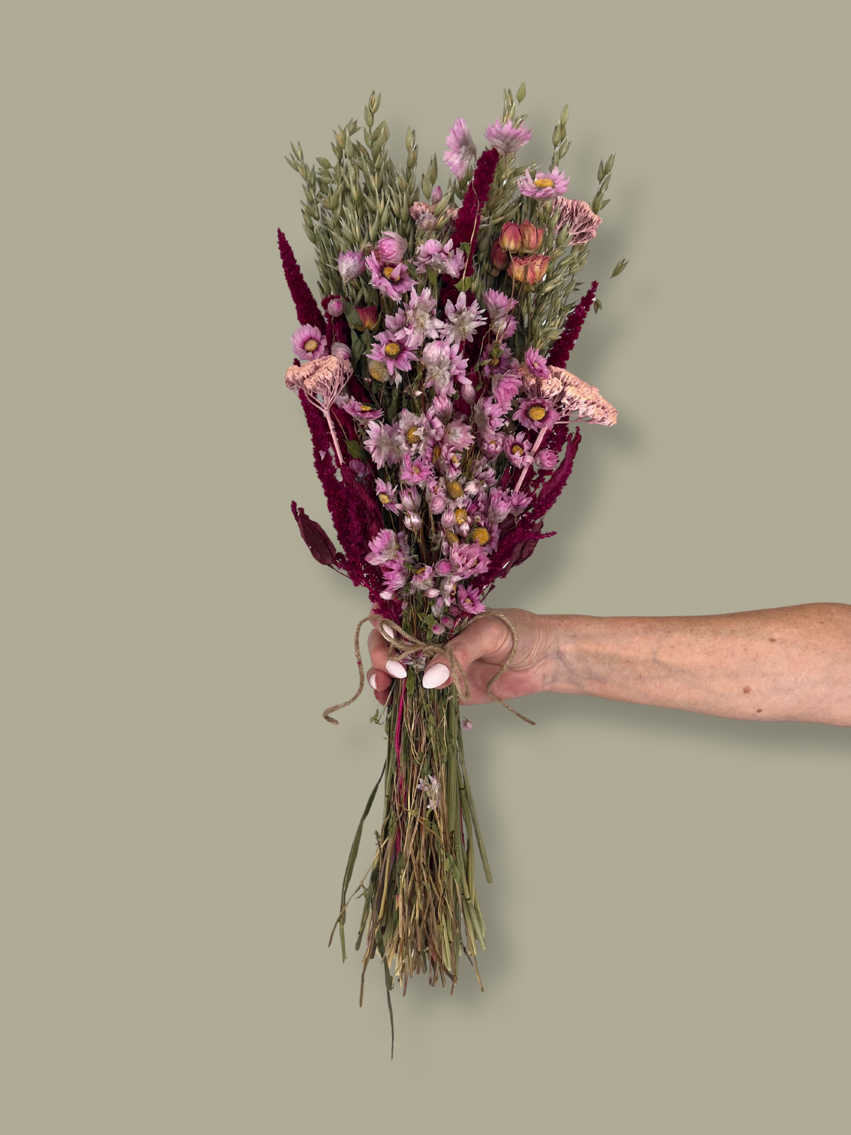 This striking, hand-tied bouquet is a harmonious blend of dried botanicals in deep red, soft pink, and natural oats. In this image a hand is holding the bouquet against a plain green background.
