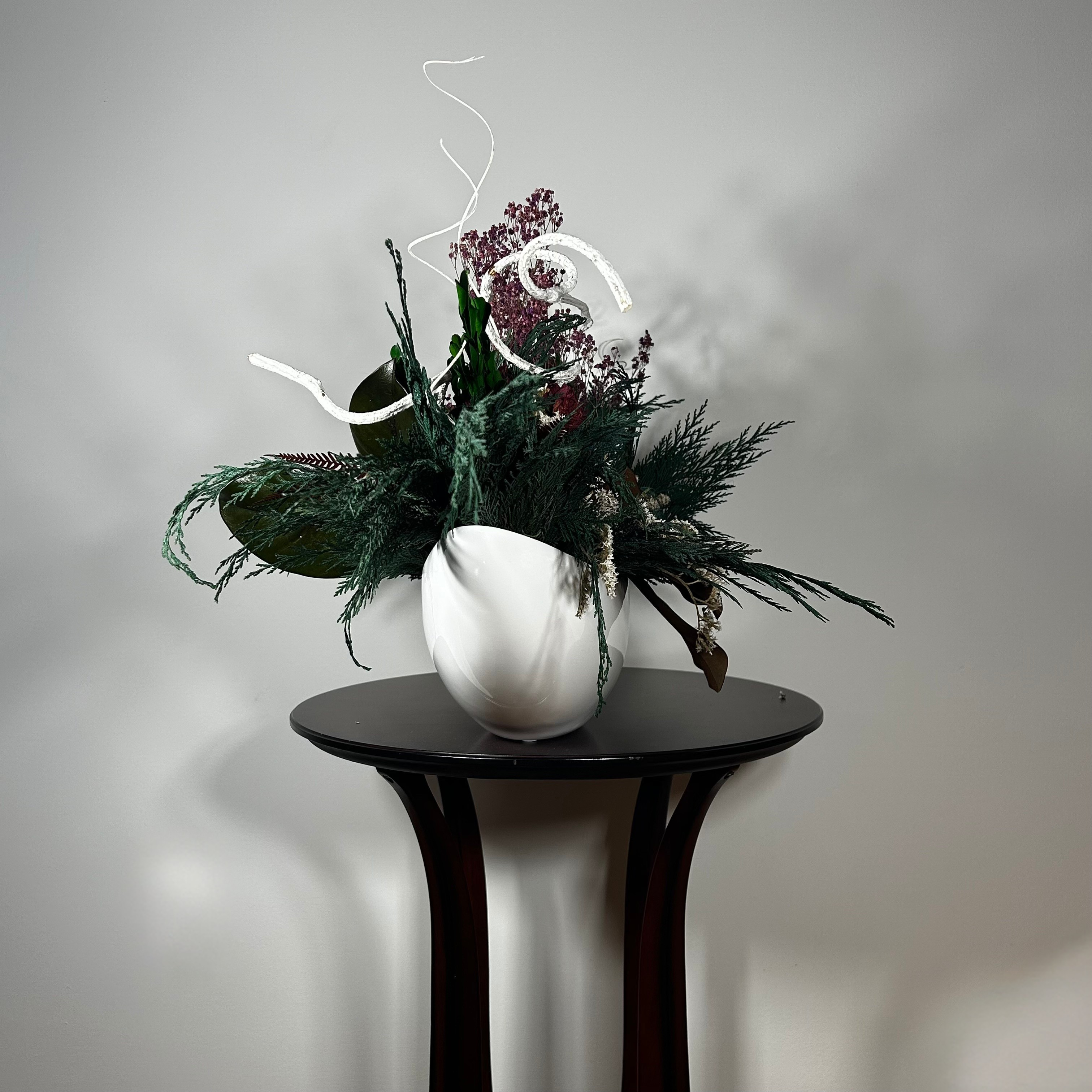 This dried flower centrepiece uses a low white lacquered vessel in a crescent shape. The foundation is a textural marvel, featuring the rich greens of preserved Cedar and Boxwood, contrasted by the striking texture of preserved Magnolia leaves and preserved burgundy Bracken. The dramatic florals include magnificent dried Dahlias in a deep crimson and elegant preserved Gypsophila dyed deep rose. Dried German Statice adds airy texture, and white curly ting ting and white wash curly vine complete the look.