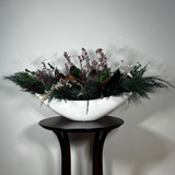 This dried flower centrepiece uses a low white lacquered vessel in a crescent shape. The foundation is a textural marvel, featuring the rich greens of preserved Cedar and Boxwood, contrasted by the striking texture of preserved Magnolia leaves and preserved burgundy Bracken. The dramatic florals include magnificent dried Dahlias in a deep crimson and elegant preserved Gypsophila dyed deep rose. Dried German Statice adds airy texture, and white curly ting ting and white wash curly vine complete the look.