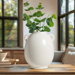 White vase with green plant on a wooden table in a room with large windows.