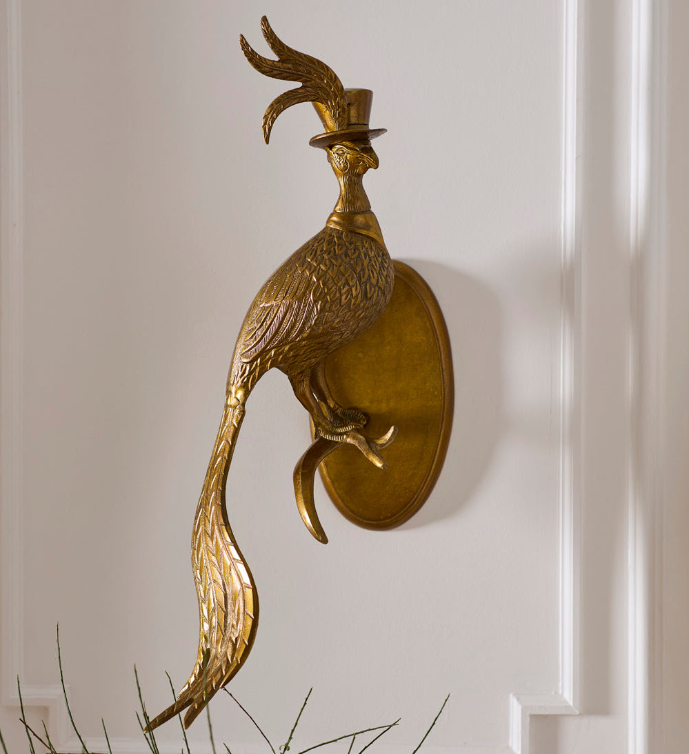 A decorative gold-toned wall sconce shaped like a peacock wearing a small top hat, mounted on a white paneled wall with a hint of greenery at the bottom.
