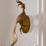 A decorative gold-toned wall sconce shaped like a peacock wearing a small top hat, mounted on a white paneled wall with a hint of greenery at the bottom.