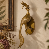 A decorative brass rooster wall sconce mounted on a textured beige wall, styled with dried flowers, books, and a framed print in a warm interior setting.