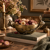 A decorative brass rabbit-shaped bowl filled with dried flowers, displayed on stacked books on a rustic wooden table with a candle and framed art in the background.