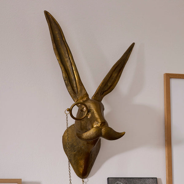 Bronze rabbit head sculpture on a wall with framed pictures and jars below.