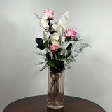 A preserved floral arrangement featuring pink roses, white flowers, and green foliage in a clear ribbed glass vase, set on a dark round table against a light gray wall.