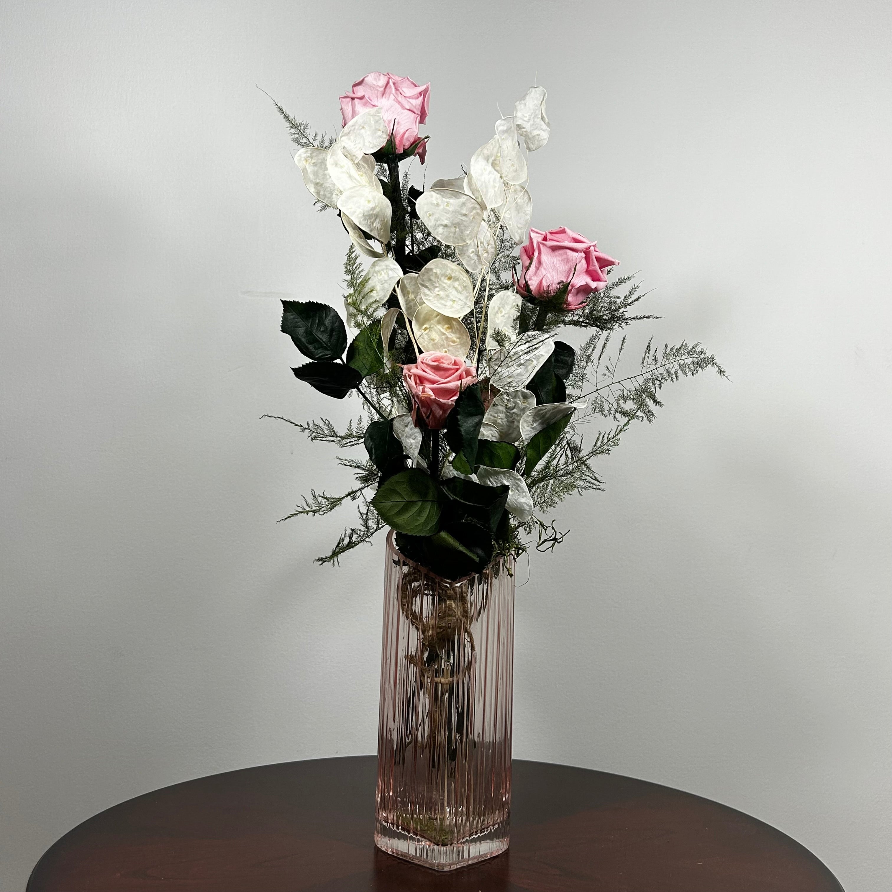A preserved floral arrangement featuring pink roses, white flowers, and green foliage in a clear ribbed glass vase, set on a dark round table against a light gray wall.