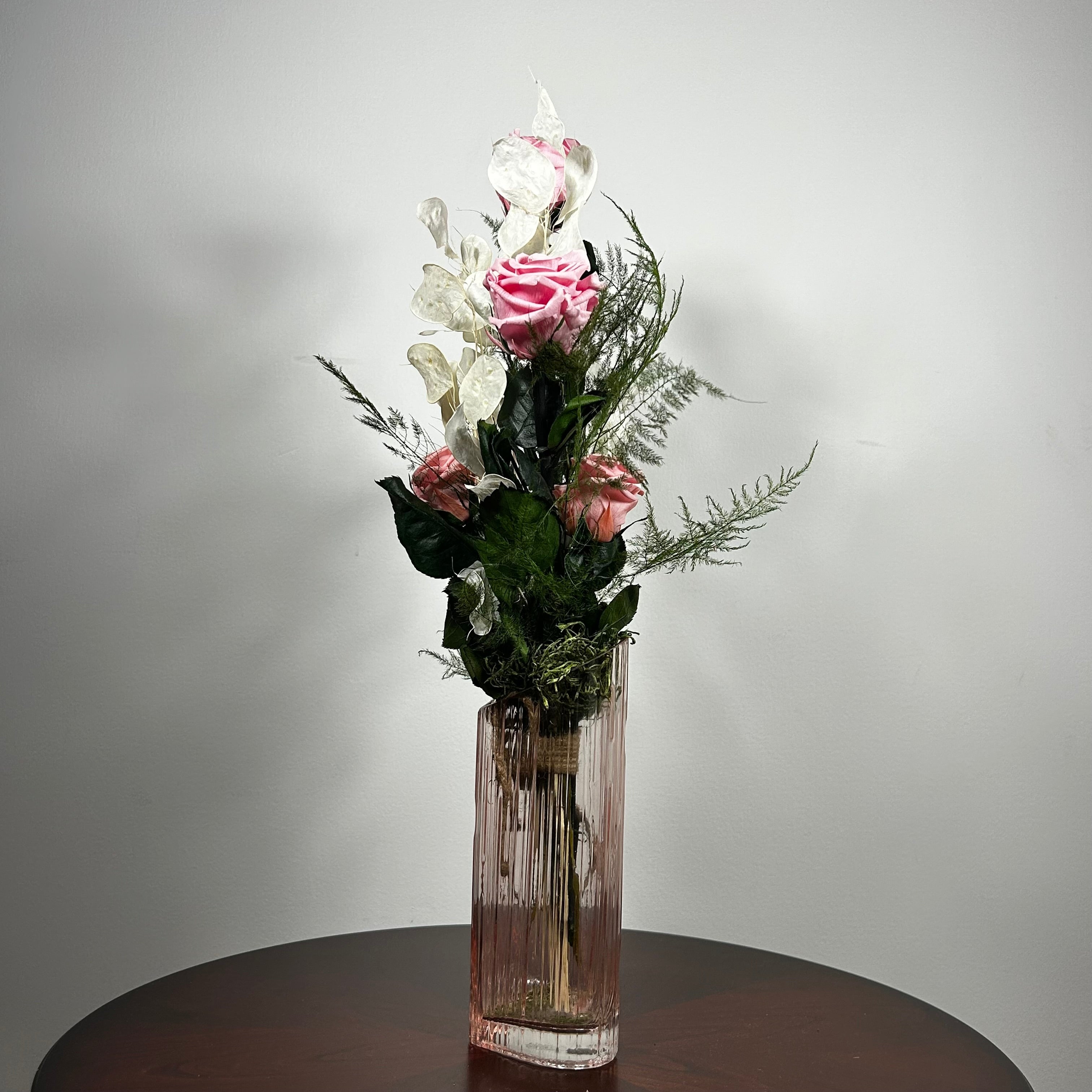 A floral arrangement featuring pink roses, white flowers, and green foliage in a tall, ribbed clear glass vase, set on a dark round table against a plain light gray wall.