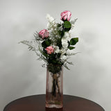 A preserved flower arrangement featuring pink roses, white blossoms, and green foliage, presented in a ribbed, clear glass vase on a dark wooden table against a neutral gray background.