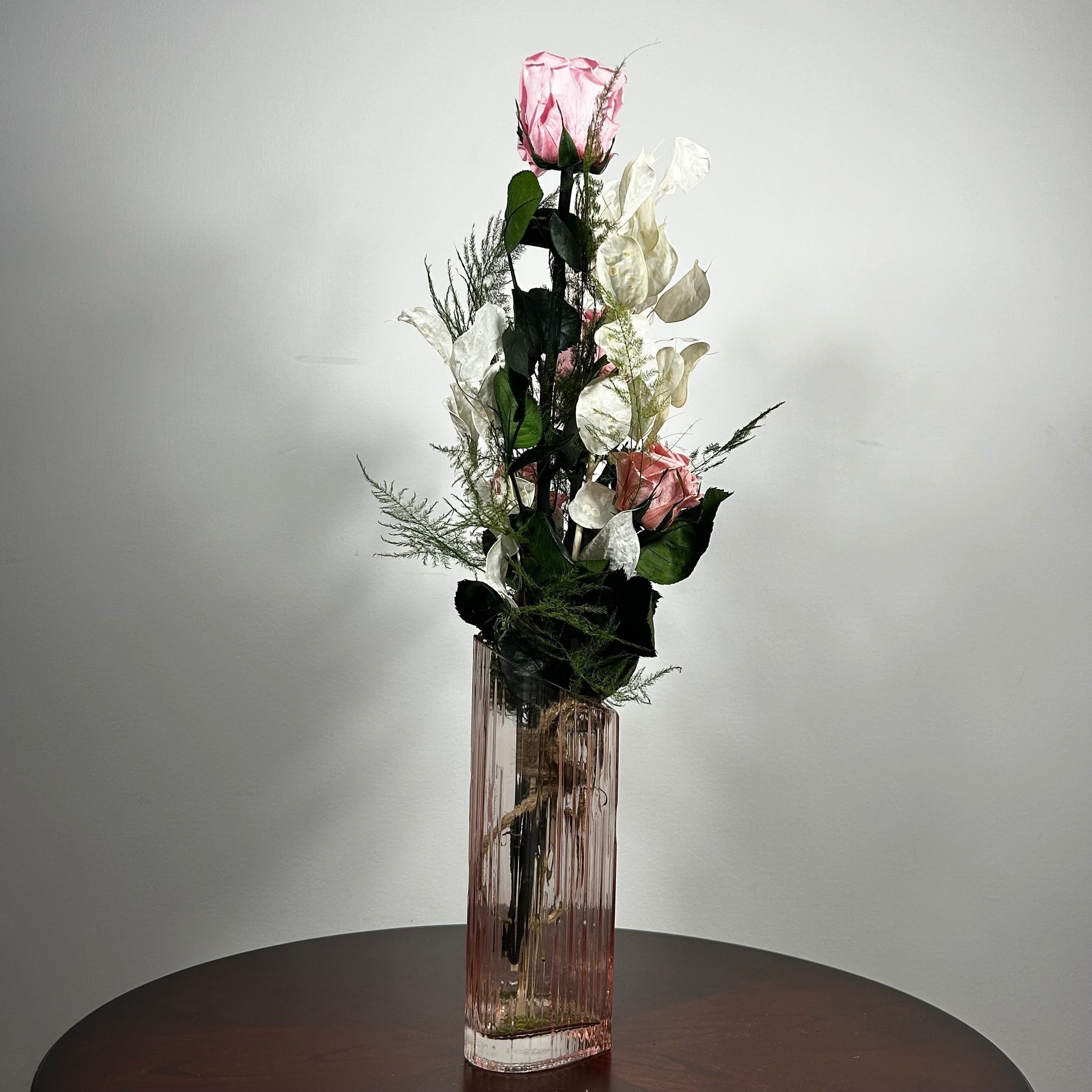 A preserved floral arrangement featuring pink roses, white blossoms, and green foliage in a tall, ribbed clear glass vase, set on a dark round table against a plain light gray wall.