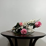 A preserved floral arrangement with pink roses and white blossoms on a dark wooden table against a plain light gray wall.