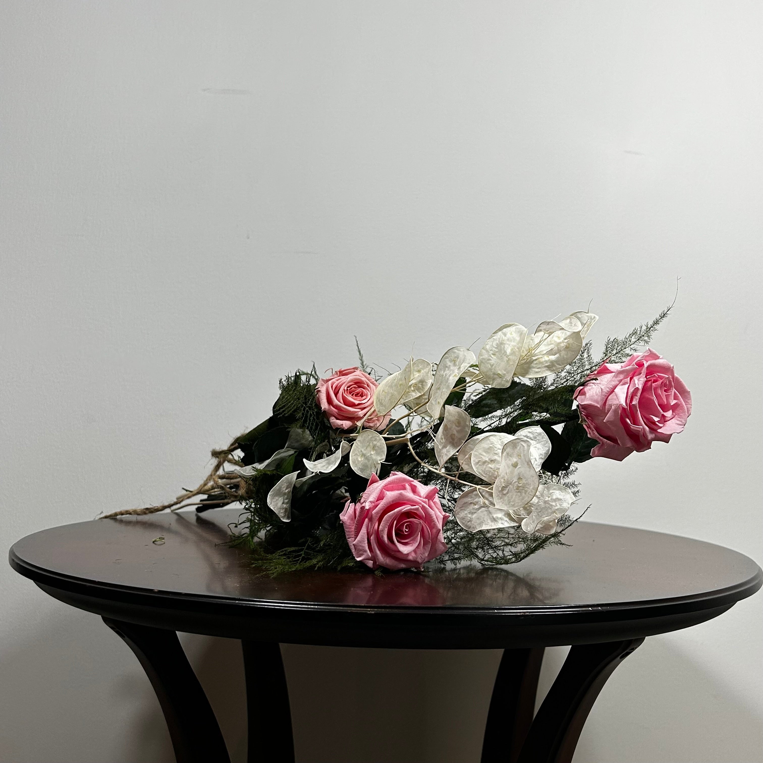 A preserved floral arrangement with pink roses and white blossoms on a dark wooden table against a plain light gray wall.