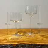 Two clear glass stemmed candle holders shaped like goblets on a wooden surface with a neutral background with height measurements. One is 9.8 inches and the other 7.8 inches