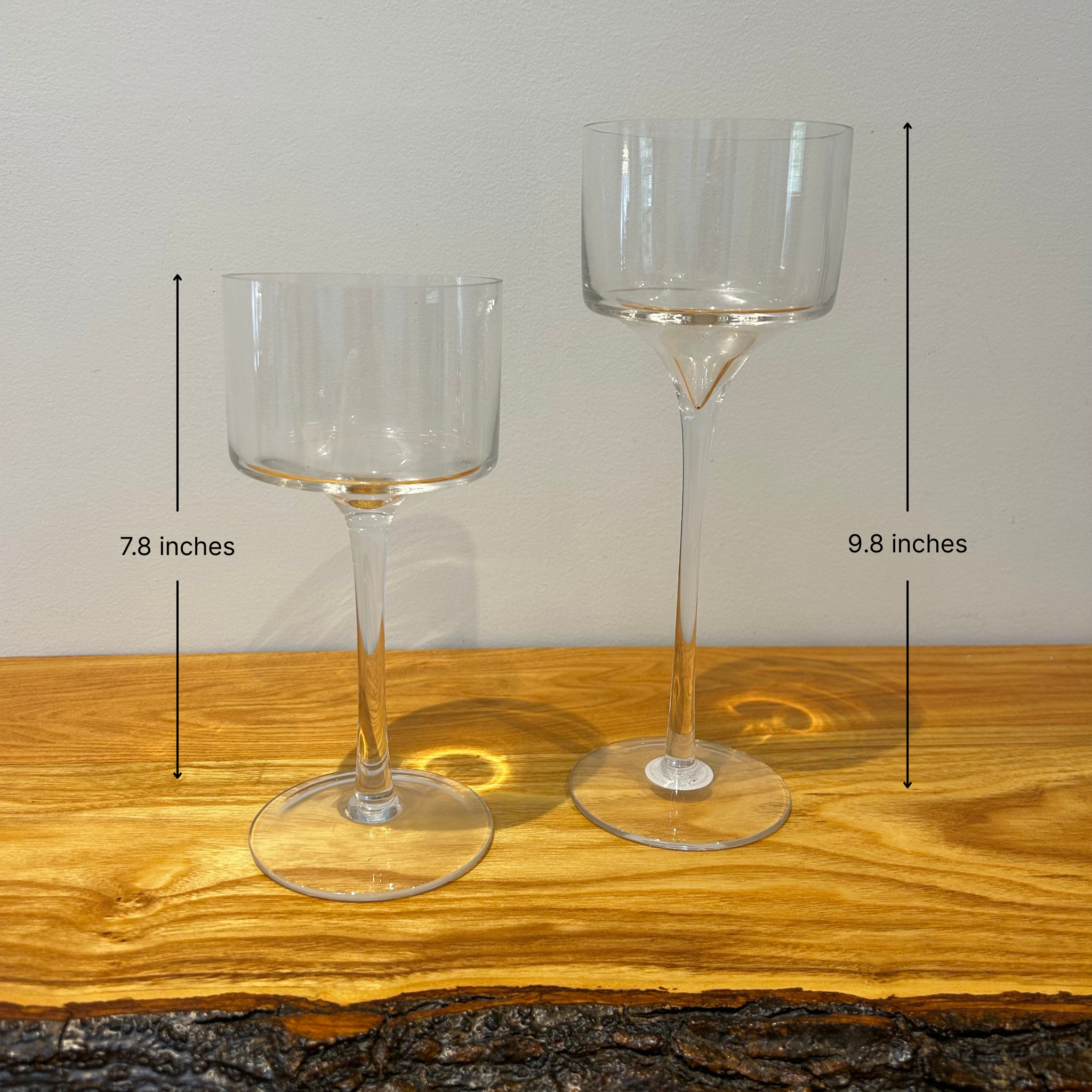 Two clear glass stemmed candle holders shaped like goblets on a wooden surface with a neutral background with height measurements. One is 9.8 inches and the other 7.8 inches