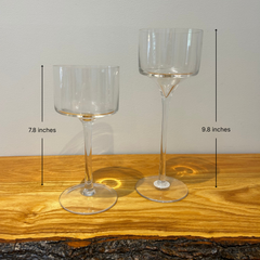 Two clear glass stemmed candle holders shaped like goblets on a wooden surface with a neutral background with height measurements. One is 9.8 inches and the other 7.8 inches
