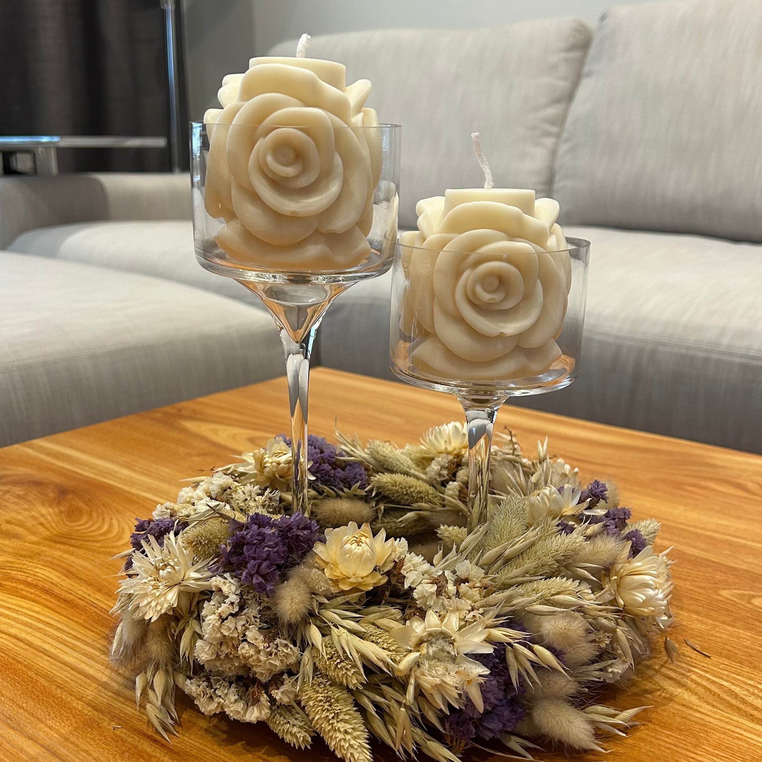 Beautiful rose-shaped soy wax candles in glass stemmed candle holders surrounded by a dried floral wreath on a wooden table.