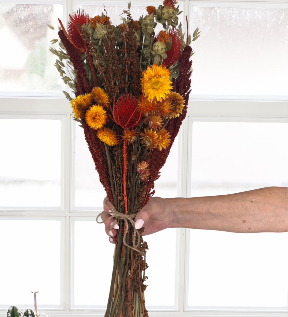 Beautifully detailed bouquet of dried flowers in Autumn colours held by a hand in front of a window with decorative items on a windowsill.