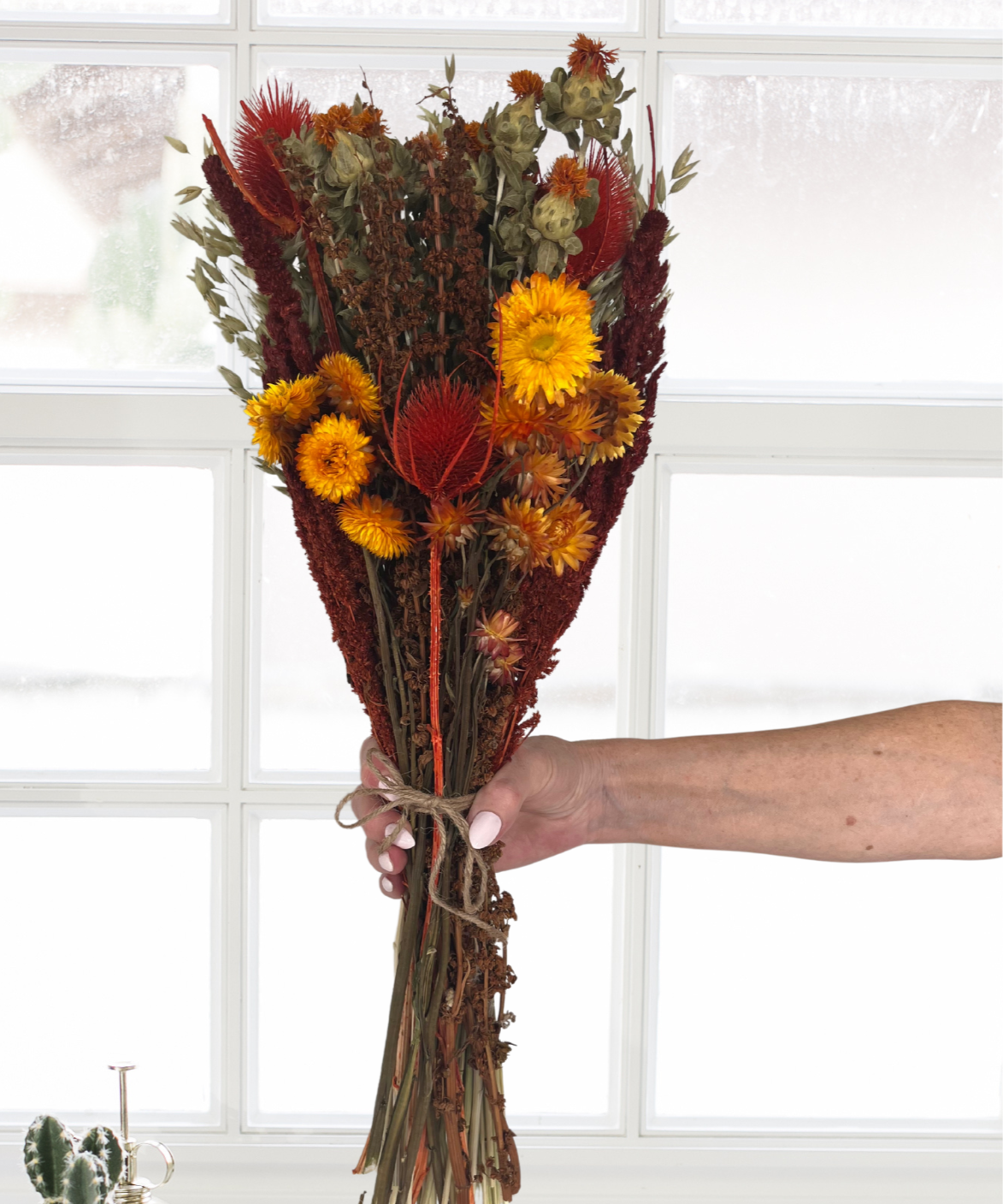 Beautifully detailed bouquet of dried flowers in Autumn colours held by a hand in front of a window with decorative items on a windowsill.
