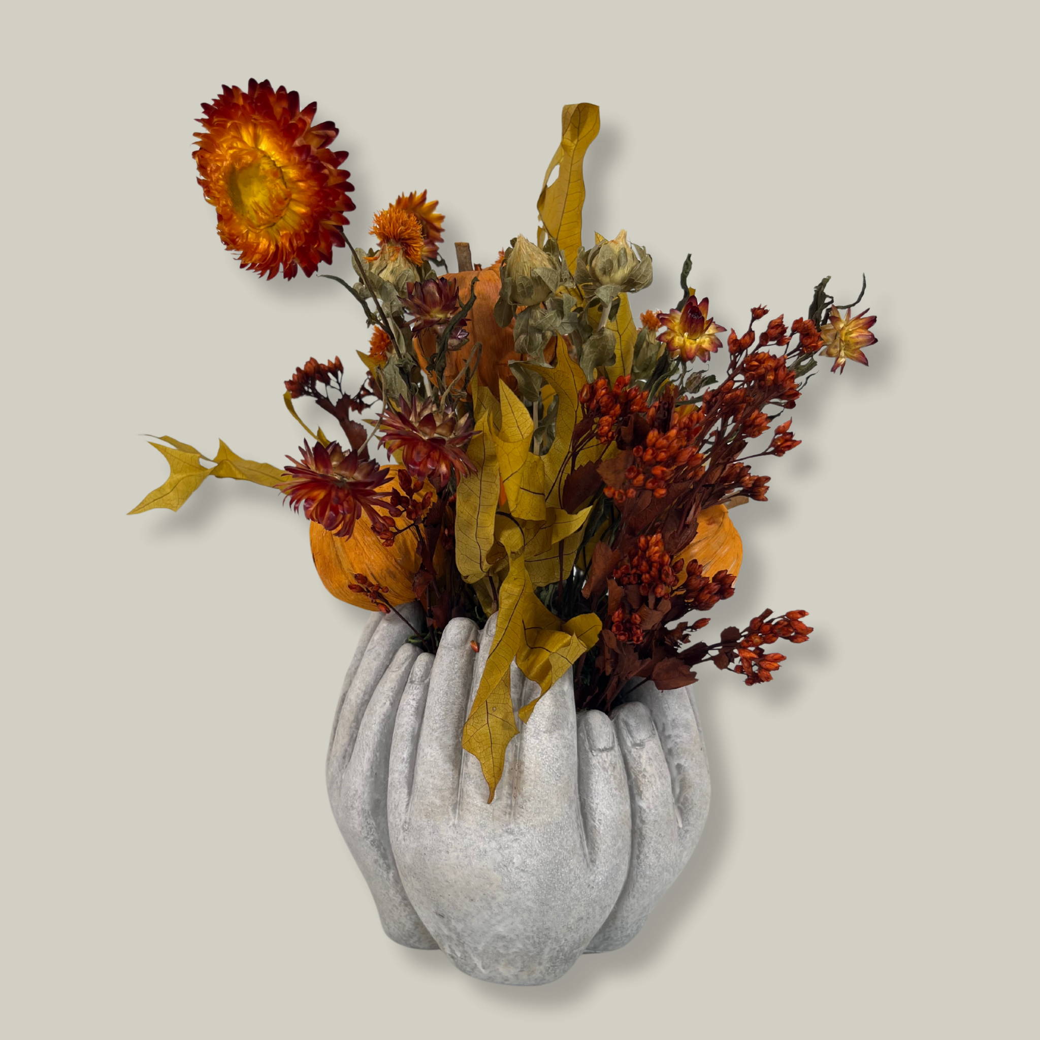 A beautiful concrete vessel sculpted to resemble for hands joined in a circle that features the bold, preserved tones of yellow scarlet oak leaves and the deep, rich crimson of Chuisco Grass. Fiery Carthamus (safflower) and luminous Helichrysums (strawflowers) in golden and orange tones add a radiant glow. The arrangement is completed with three small pumpkins, perfectly nestled within the blooms.