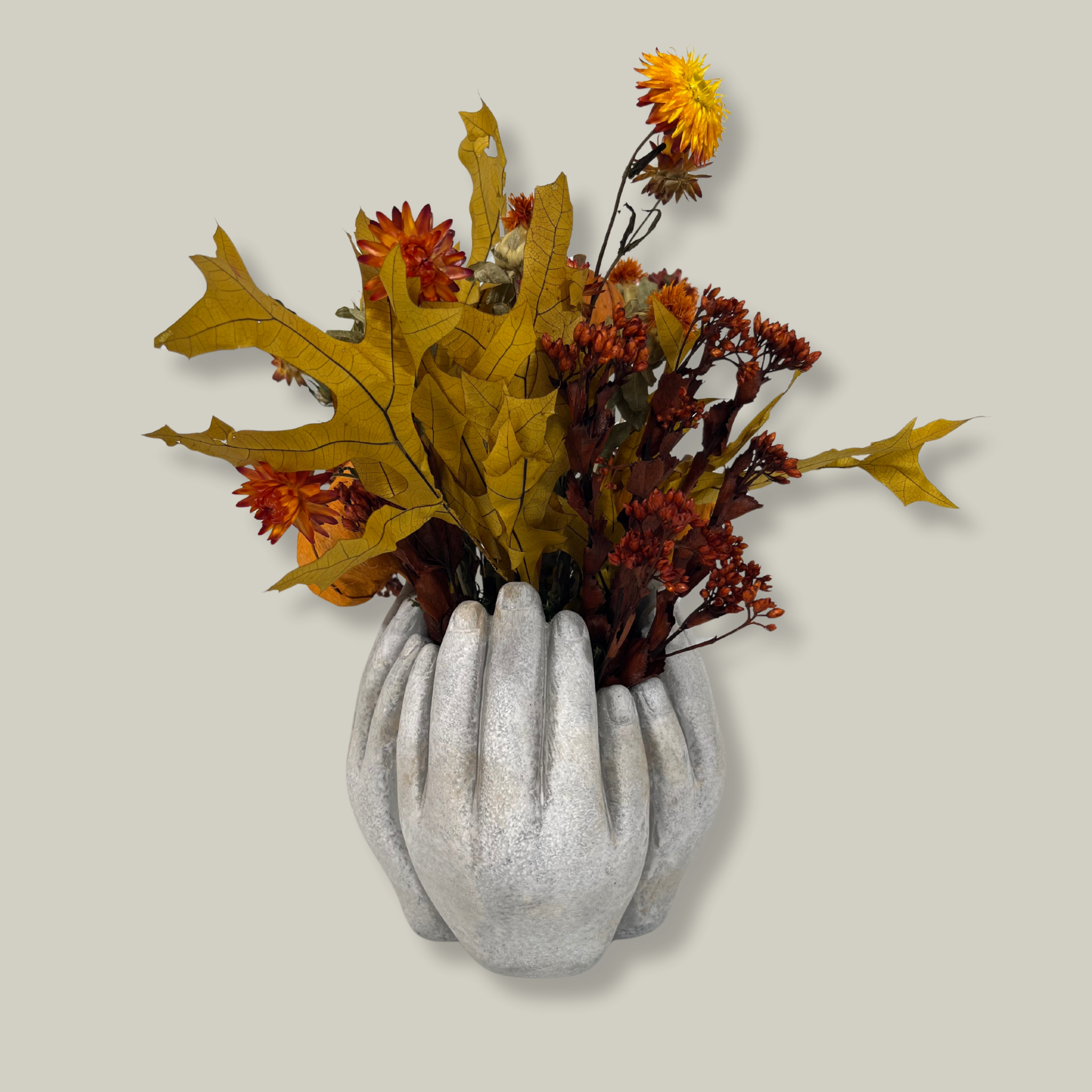 A beautiful concrete vessel sculpted to resemble for hands joined in a circle that features the bold, preserved tones of yellow scarlet oak leaves and the deep, rich crimson of Chuisco Grass. Fiery Carthamus (safflower) and luminous Helichrysums (strawflowers) in golden and orange tones add a radiant glow. The arrangement is completed with three small pumpkins, perfectly nestled within the blooms.