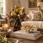 A styled interior scene with a dark amber glass vase holding a dried floral arrangement in a living room. The arrangement includes yellow, cream, and brown botanicals.