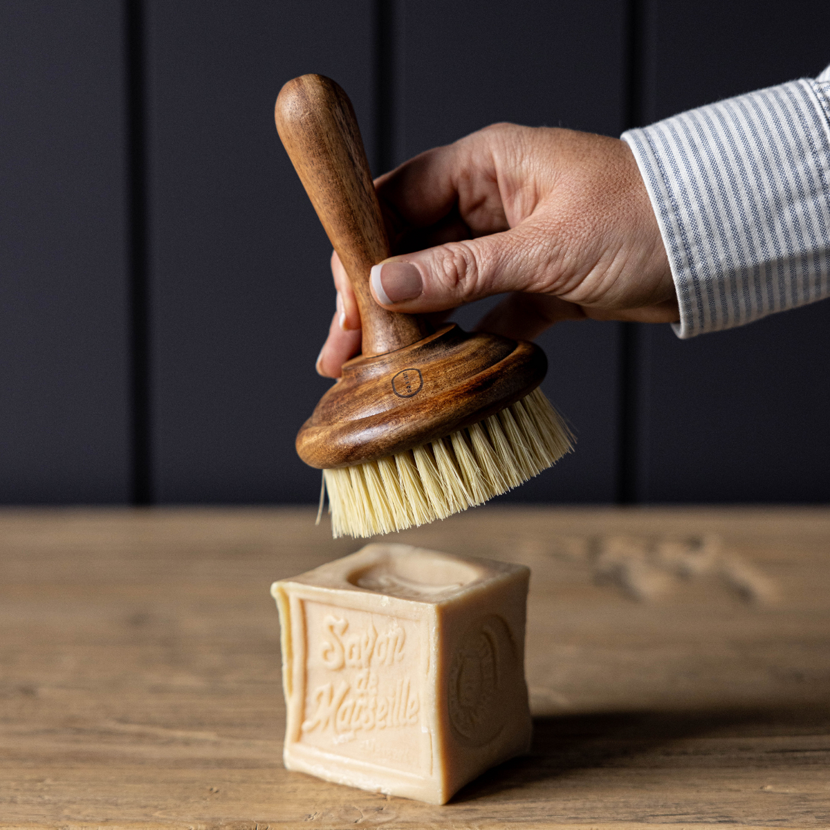 Hand holding a wooden dish brush over a block of soap on a wooden surface with a dark background