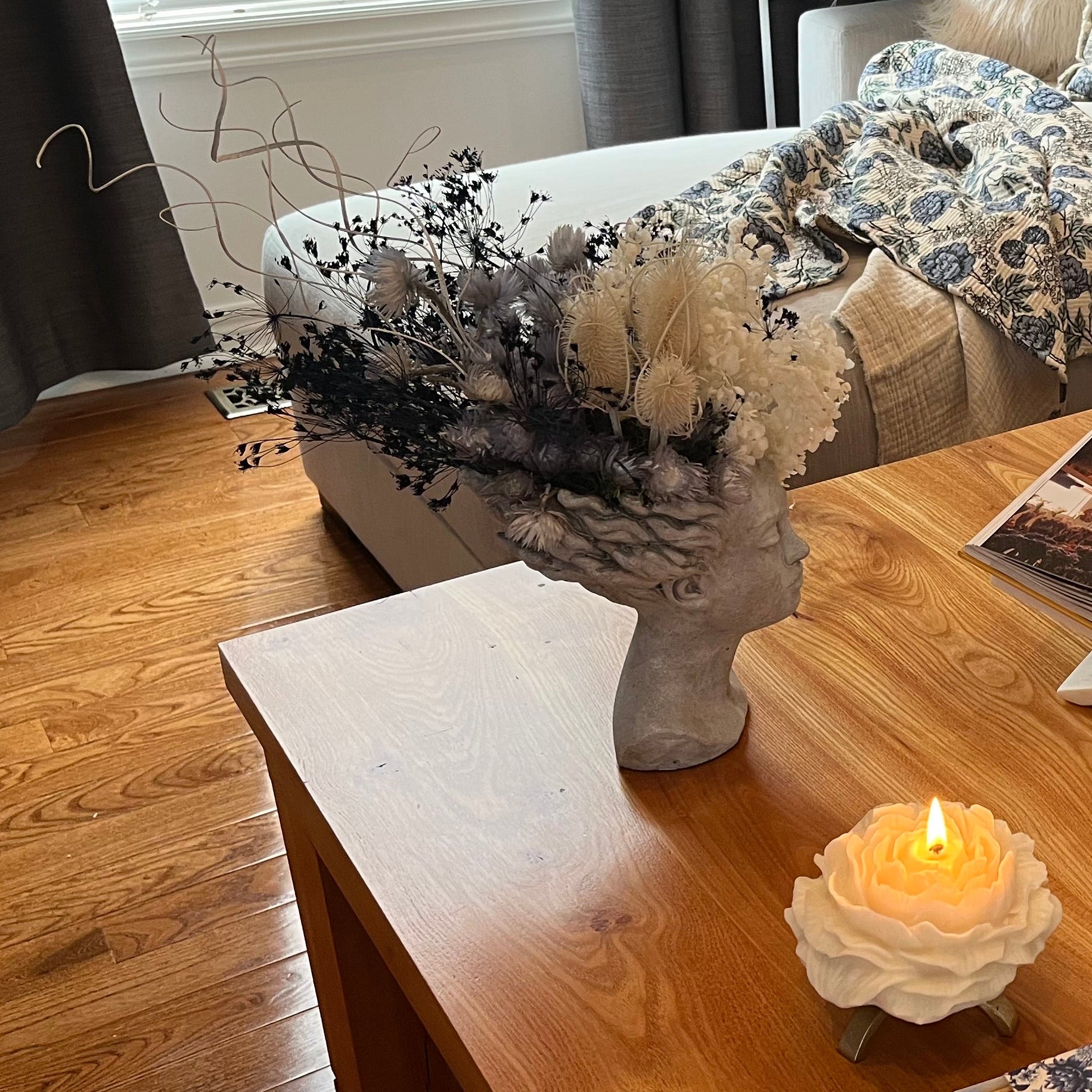 This concrete vessel, beautifully sculpted as a woman's serene face, her hair appears to be caught in a soft breeze, with a dried floral arrangement made up navy bupleurum , white curly ting ting, delicate light blue cape flowers, a cluster of three bleached teasels adorns the front left side of her face, and a pure white hydrangea on the right. The arrangement is on a wooden coffee table set in a living room.