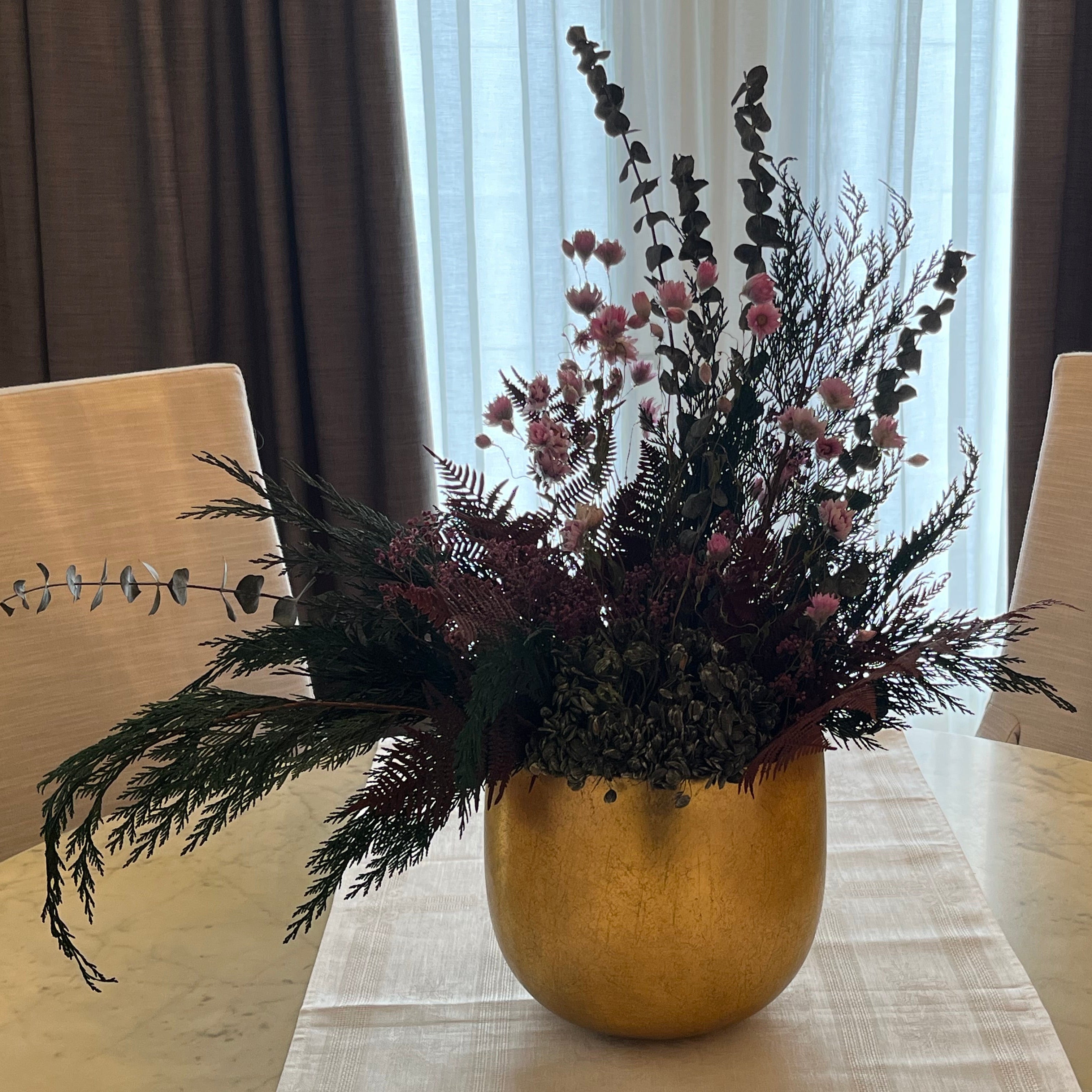 Gold vase with dried flowers on a table with chairs and curtains in the background