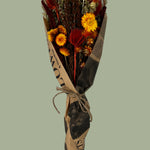 Bouquet titled "Autumn Ember Bouquet" is designed to be a magnificent focal point, perfect for a large vase on a dining table, an entryway, or a mantelpiece. In this photo it is wrapped in bouquet paper that looks like vintage newspaper against a plain green background.