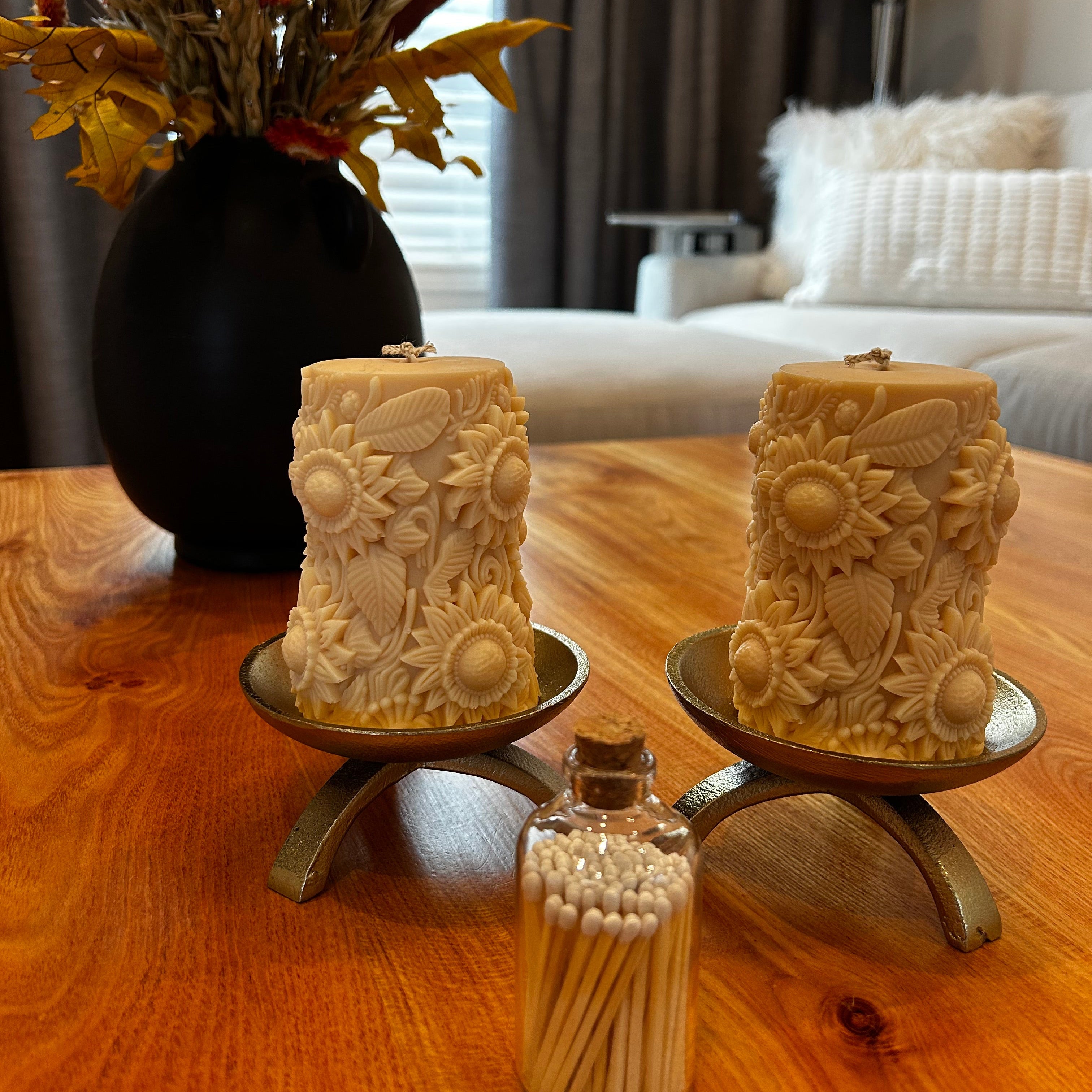 Beautiful soy wax candles embossed with sunflower motif on gold tripod candle holders atop a wooden coffee table. There is also a dried flower arrangement, and a jar with wooden matches displayed on the table.