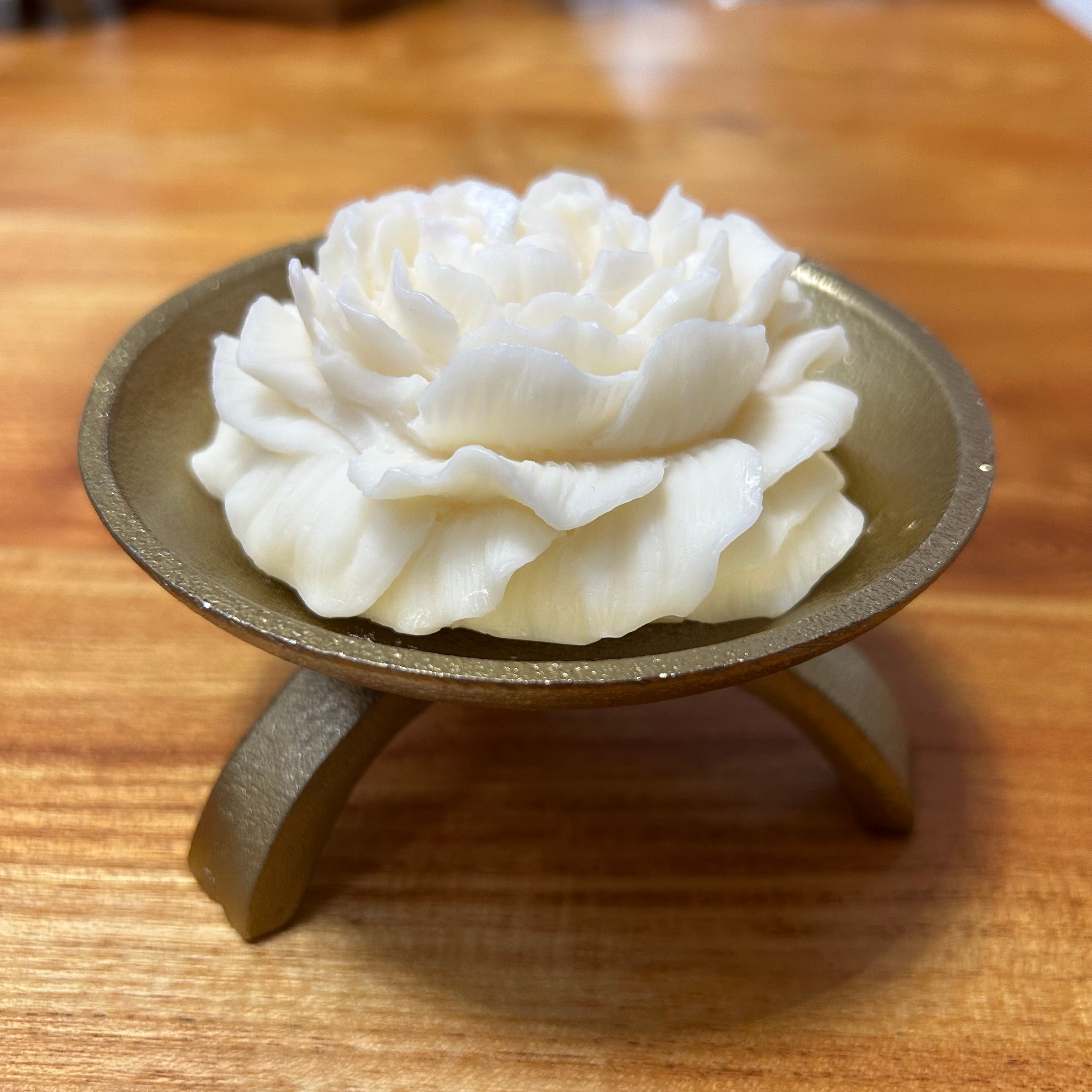 Beautiful small candle in the shape of a peony flower on a gold tripod candle holder placed on a wooden surface. 