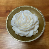 Beautiful small candle in the shape of a peony flower on a gold tripod candle holder placed on a wooden surface. 