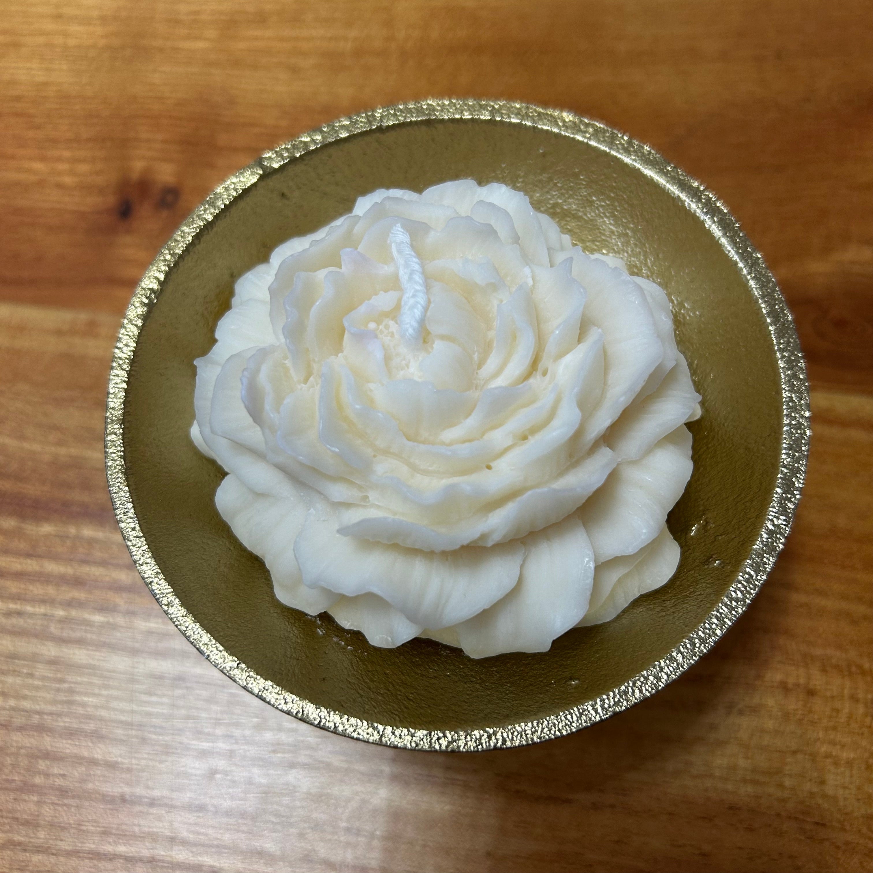 Beautiful small candle in the shape of a peony flower on a gold tripod candle holder placed on a wooden surface. 