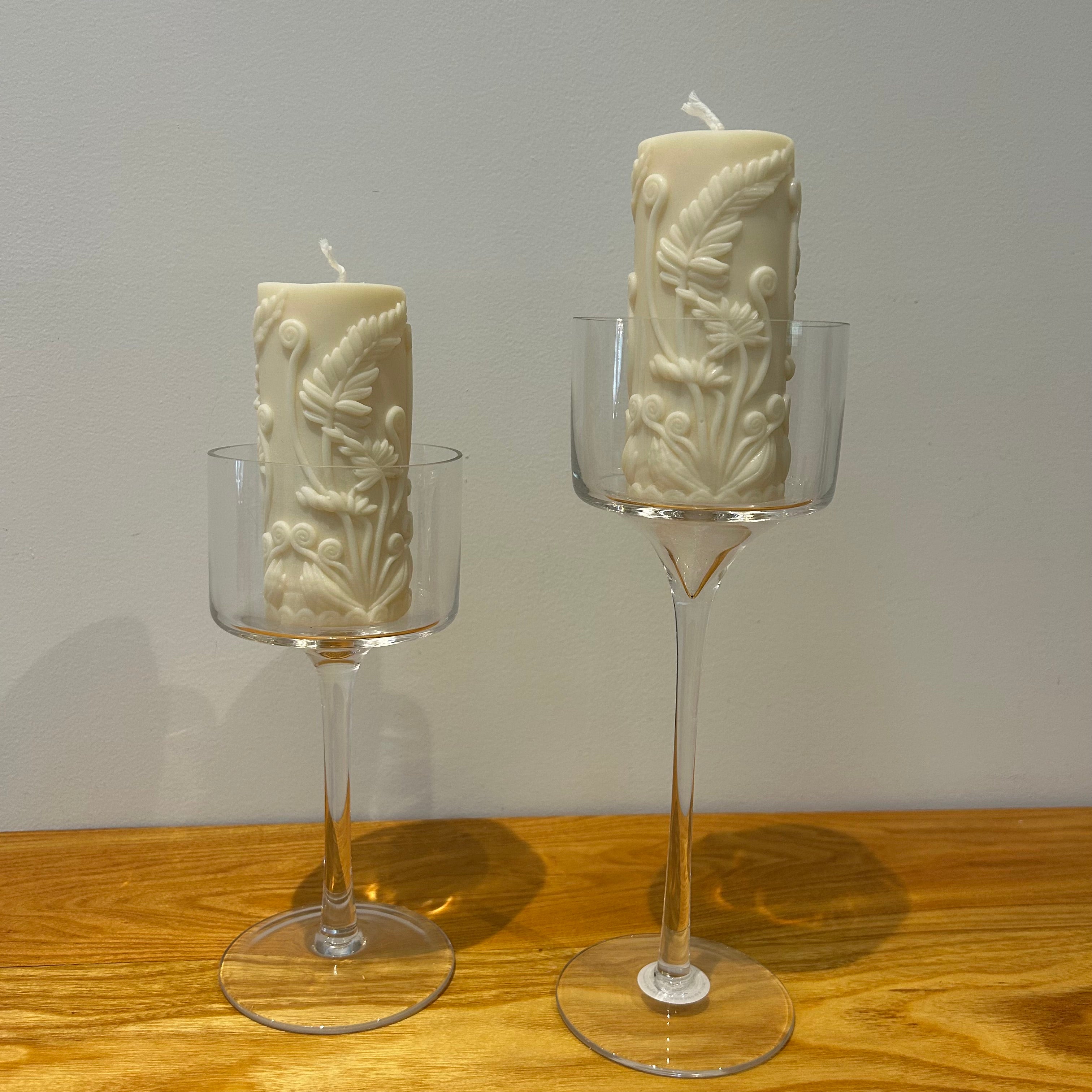 Two clear glass stemmed candle holders shaped like goblets holding pillar candles with a embossed fern motif on a wooden surface with a neutral background