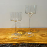 Two clear glass stemmed candle holders shaped like goblets on a wooden surface with a neutral background