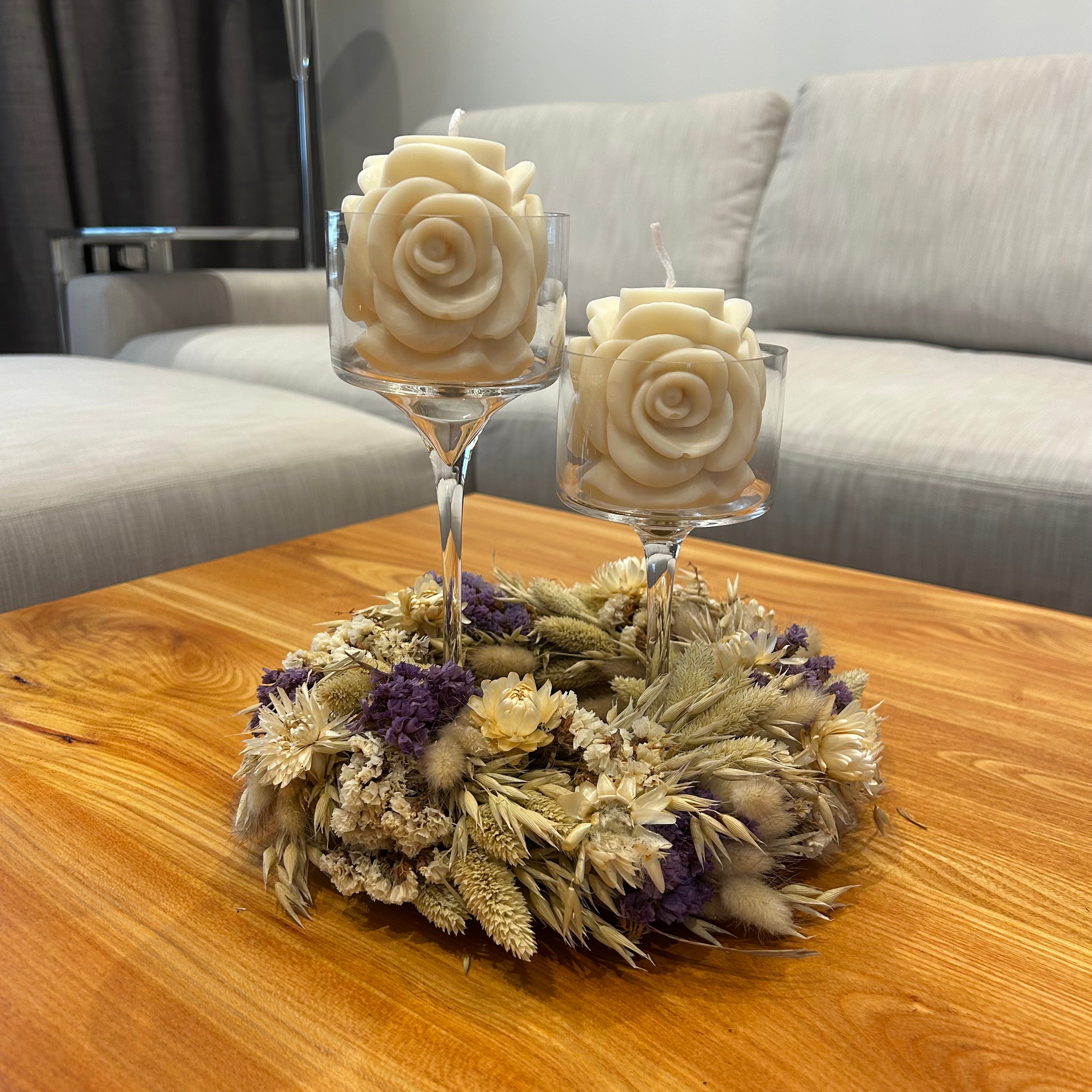 Beautiful rose-shaped soy wax candles in glass stemmed candle holders surrounded by a dried floral wreath on a wooden table.
