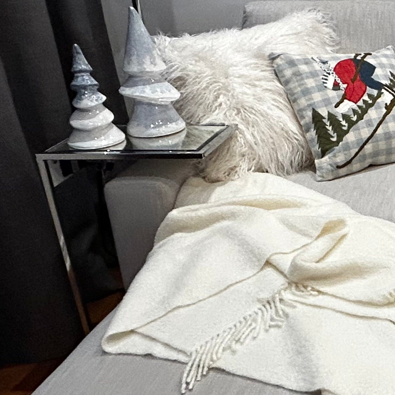Grey sofa with a ivory wool blend throw, decorative pillows, and small Christmas trees on a side table.