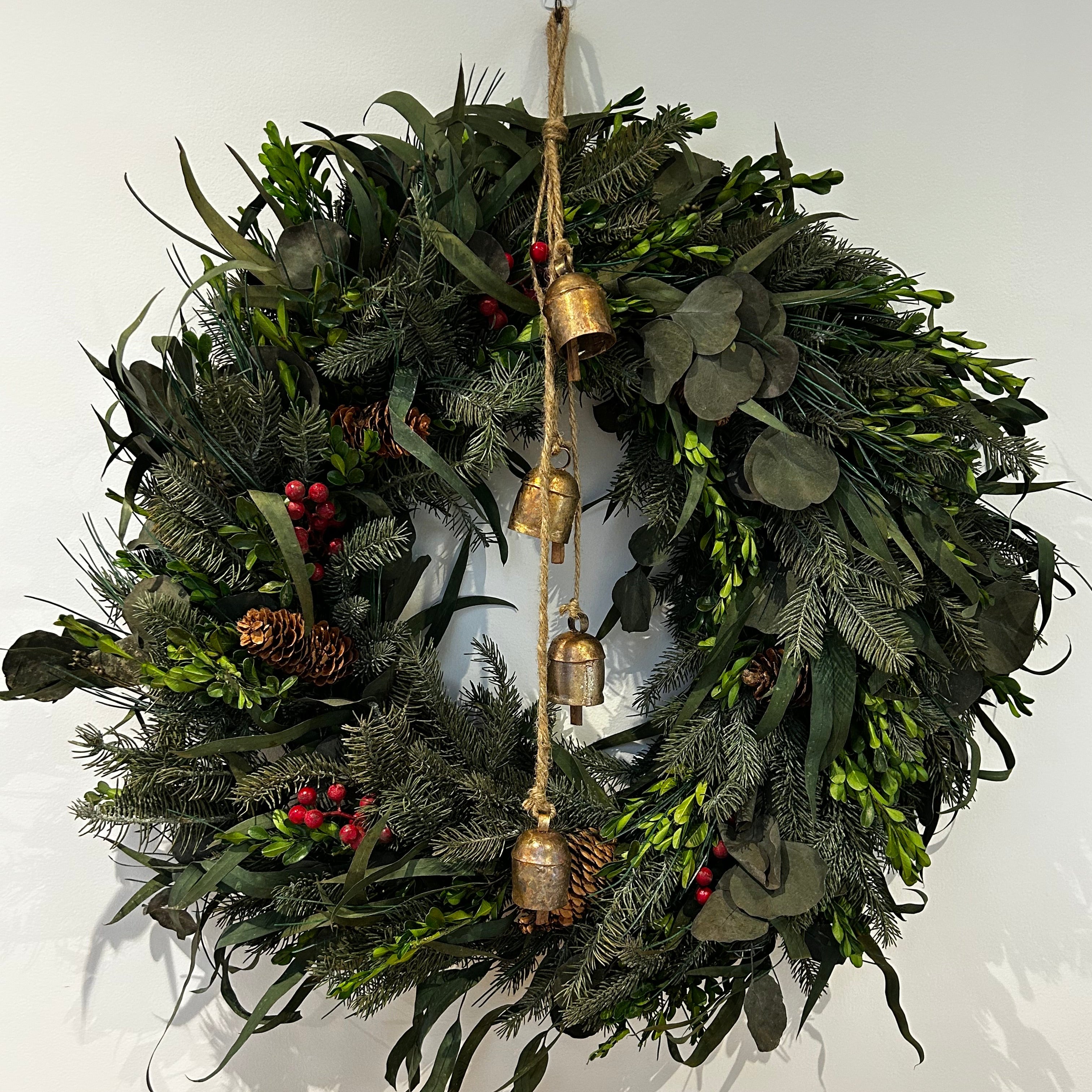 A wreath featuring a stunning foundation of deep green preserved spruce and preserved boxwood, beautifully layered with the modern forms of dried green curly eucalyptus and large round leaf eucalyptus. Traditional accents of red berries and natural pinecones are nestled throughout, completing this breathtaking tapestry of texture and colour. Accessorized with a beautiful set of hand-made iron bells with a bronze finish hanging on jute twine.
