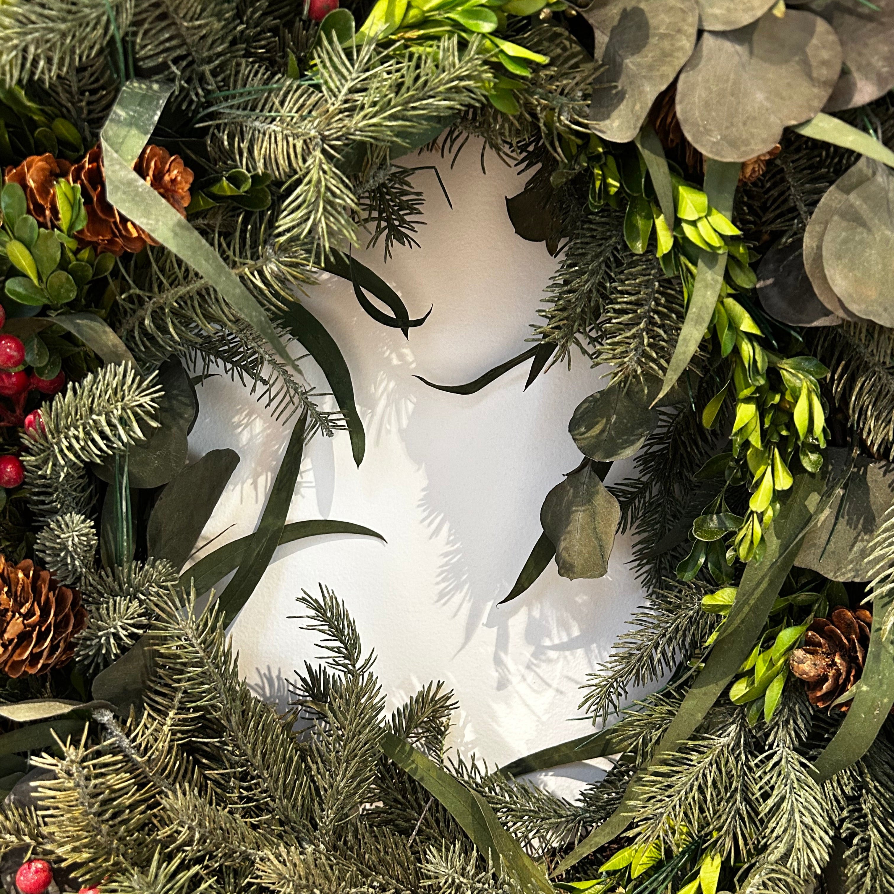 A wreath featuring a stunning foundation of deep green preserved spruce and preserved boxwood, beautifully layered with the modern forms of dried green curly eucalyptus and large round leaf eucalyptus. Traditional accents of red berries and natural pinecones are nestled throughout, completing this breathtaking tapestry of texture and colour.