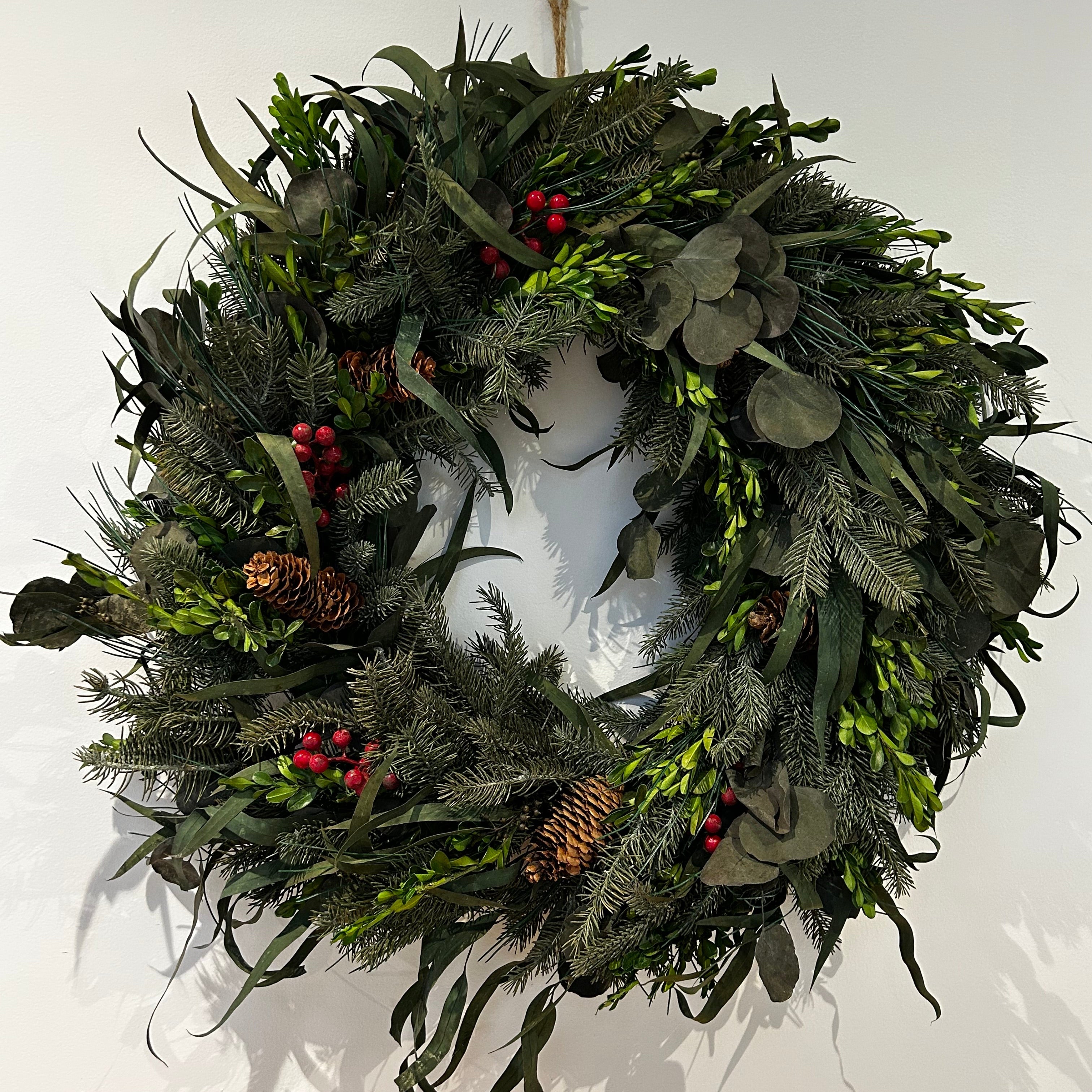 A wreath featuring a stunning foundation of deep green preserved spruce and preserved boxwood, beautifully layered with the modern forms of dried green curly eucalyptus and large round leaf eucalyptus. Traditional accents of red berries and natural pinecones are nestled throughout, completing this breathtaking tapestry of texture and colour.
