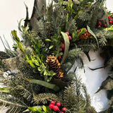 A close-up view of one side of a wreath featuring a stunning foundation of deep green preserved spruce and preserved boxwood, beautifully layered with the modern forms of dried green curly eucalyptus and large round leaf eucalyptus. Traditional accents of red berries and natural pinecones are nestled throughout, completing this breathtaking tapestry of texture and colour.
