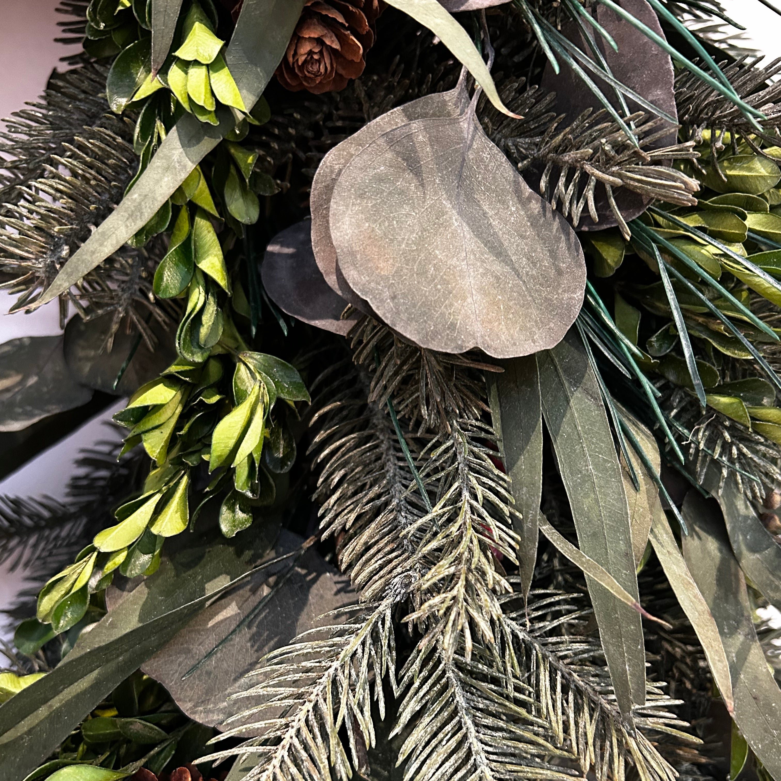 A close-up view of a wreath featuring a stunning foundation of deep green preserved spruce and preserved boxwood, beautifully layered with the modern forms of dried green curly eucalyptus and large round leaf eucalyptus. 