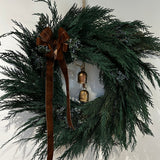 A wreath layered with lush, preserved Cedar and accented with vibrant Juniper Berries, a chocolate brown velvet bow with ribbon ends dangling, and a pair of iron bells on jute twine hanging in the middle.