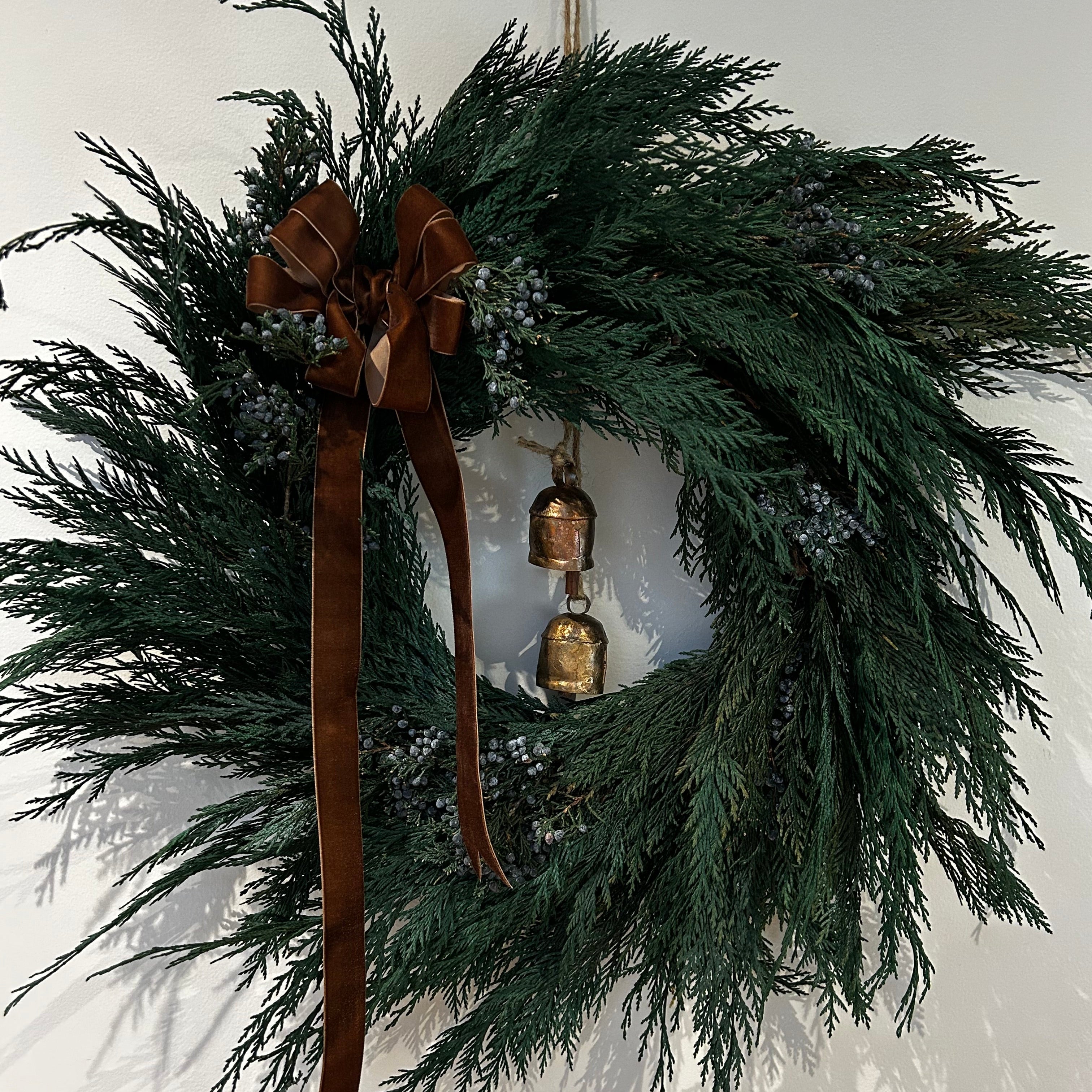 A wreath layered with lush, preserved Cedar and accented with vibrant Juniper Berries, a chocolate brown velvet bow with ribbon ends dangling, and a pair of iron bells on jute twine hanging in the middle.